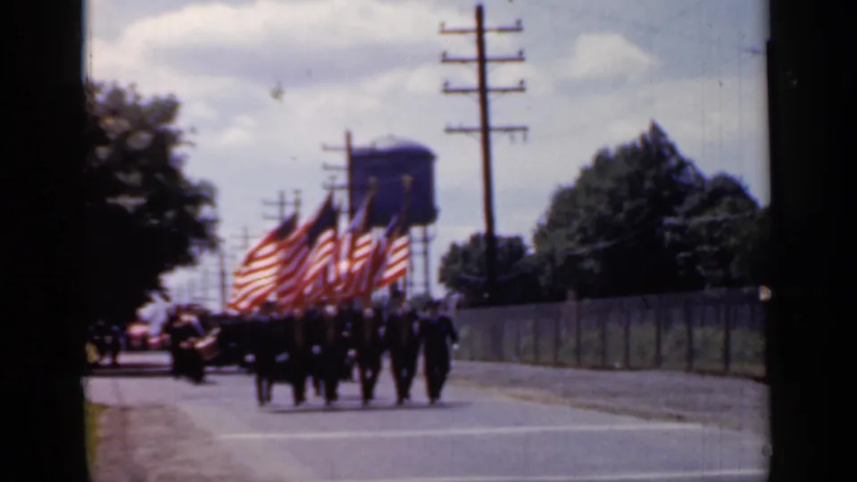 A flag-bearing parade unit marches along a Mineola street in 1966, with a water tower and utility poles in the background.
