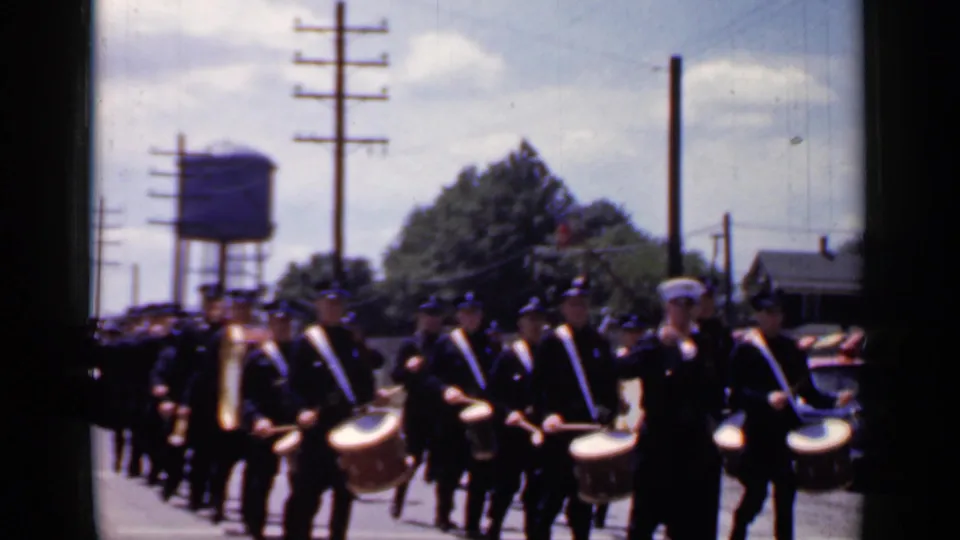 Uniformed marching band members with drums march in the Mineola parade, with a water tower and utility poles behind them.