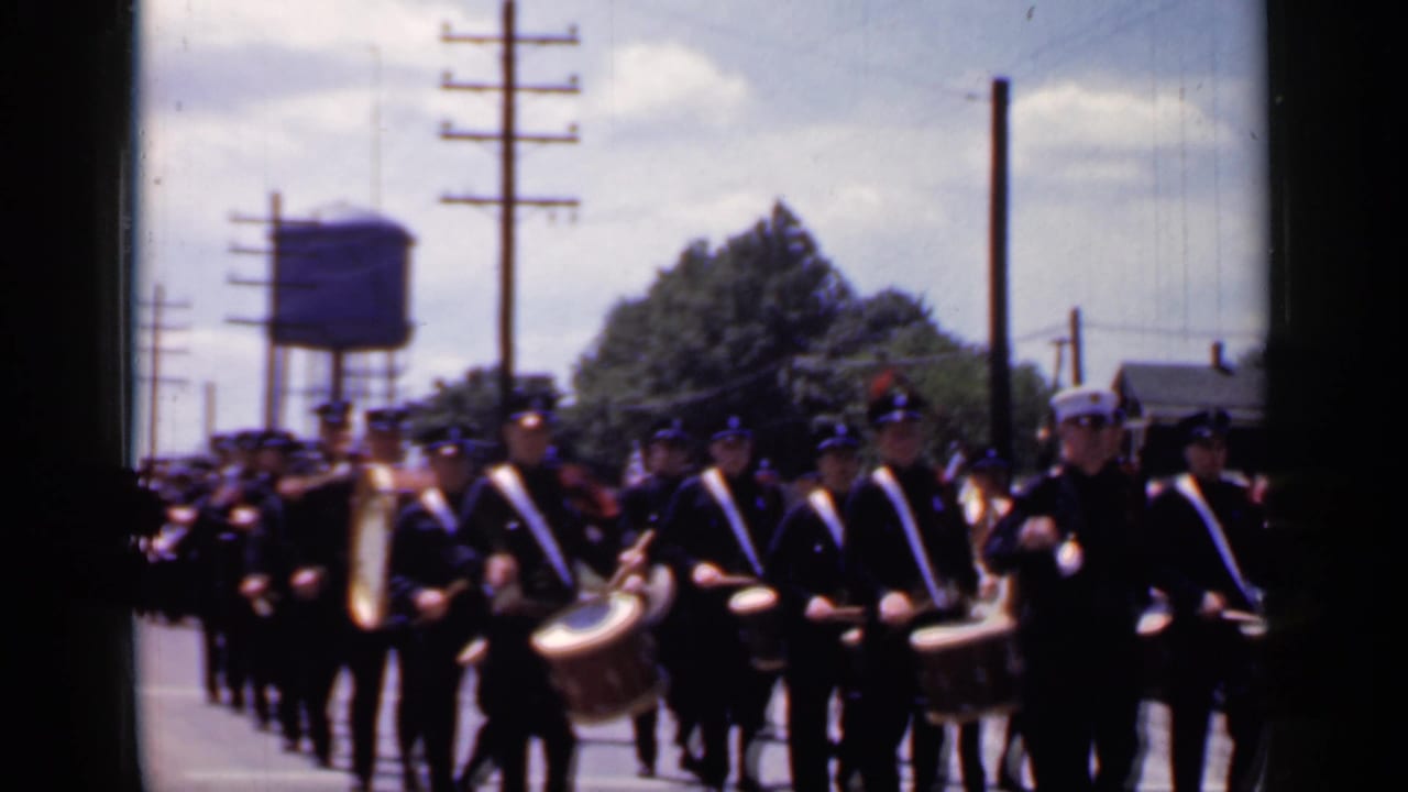 Uniformed parade band and marchers on a sunny street with utility poles and a water tower.