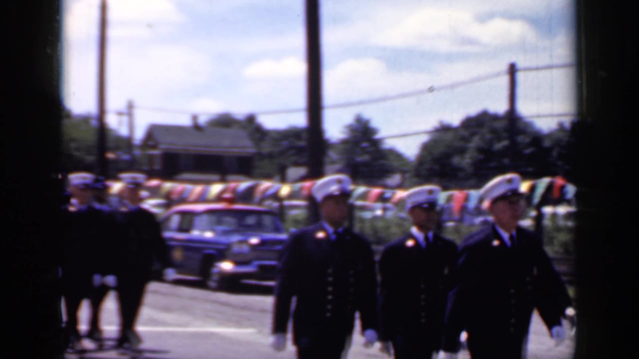 Uniformed participants in a 1966 Mineola parade pass vintage cars and roadside bunting beneath overhead utility wires.