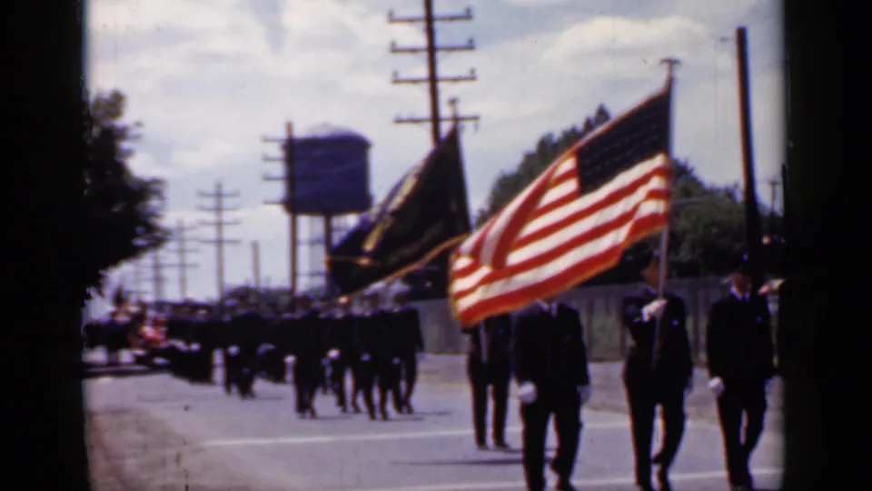 1966 Mineola parade view with an American flag, a dark flag, uniformed marchers, utility poles, and a water tower in the background.