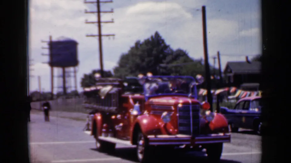A red vintage fire truck proceeds through the 1966 Mineola parade, with a water tower, parked cars, flags, and utility poles behind it.