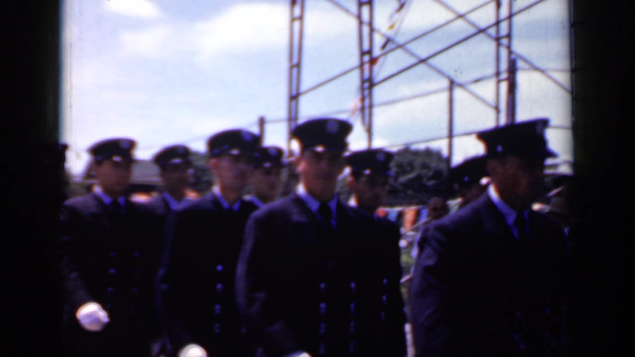 In Mineola, New York in 1966, uniformed men in dark double-breasted jackets and peaked caps march in a street parade.