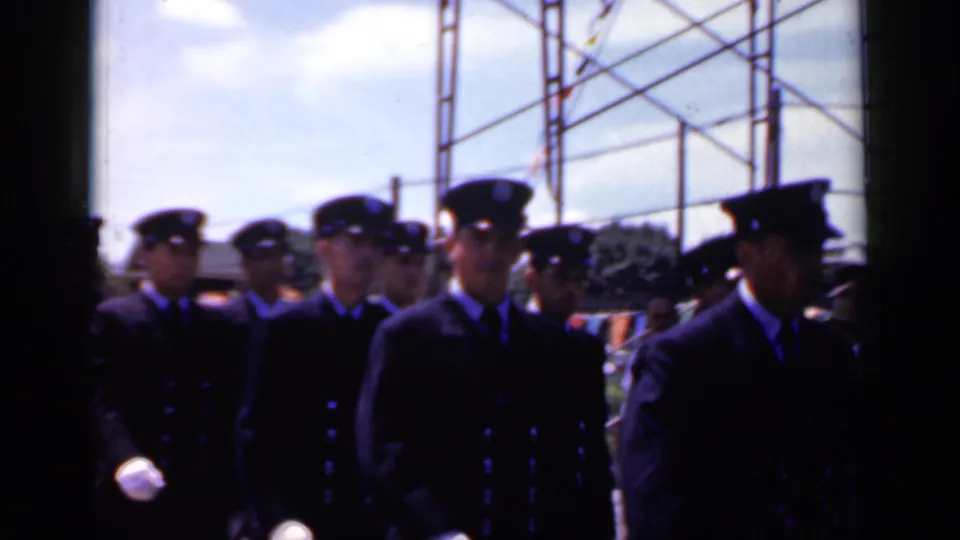 In Mineola, New York in 1966, uniformed men in dark double-breasted jackets and peaked caps march in a street parade.