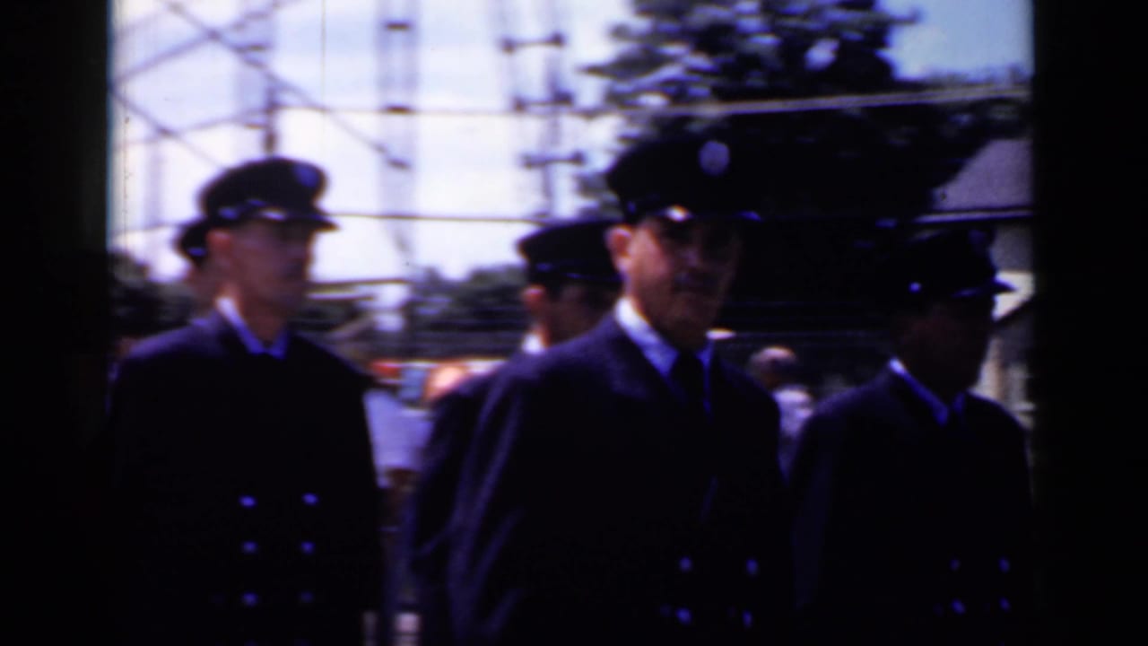 Men in dark uniforms continue marching past poles and roadside decorations.