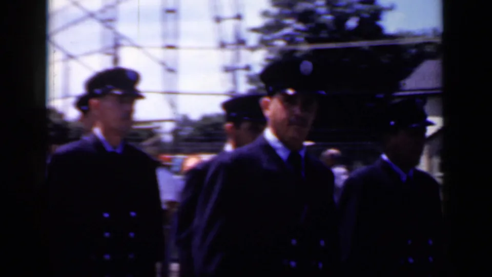 A line of uniformed men marches through the Mineola parade route, with utility poles and colorful decorations in the background.
