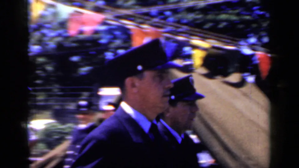 A closer 1966 Mineola parade view shows men in peaked caps and dark uniforms moving past colorful overhead bunting.