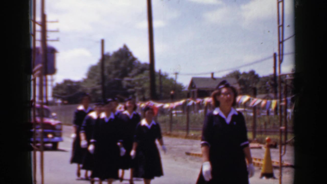 Uniformed women walk toward the camera on a sunlit street beside pennant flags and utility poles.