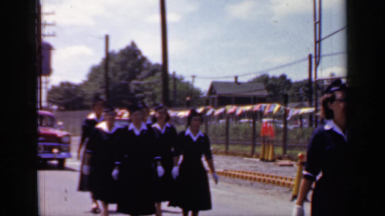 A line of women in matching dark uniforms marches past a fence with colorful pennants behind them.