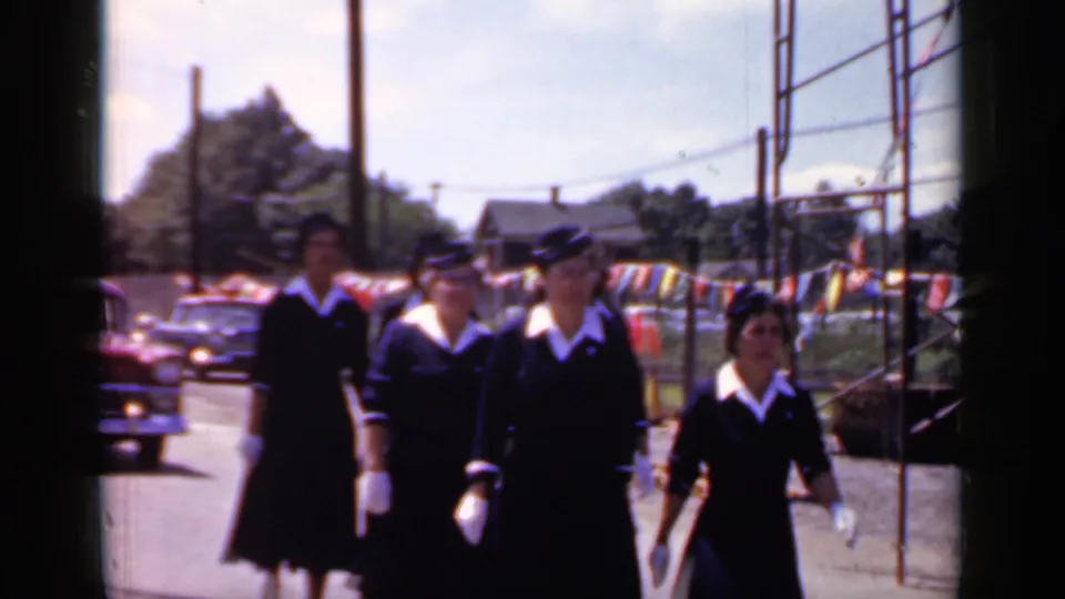 At the end of the clip, uniformed women march past the camera in Mineola, New York, with a red car and fairground-like pennants behind them.