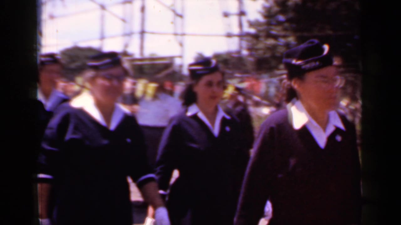 Uniformed women march past a curbside camera during a neighborhood parade.