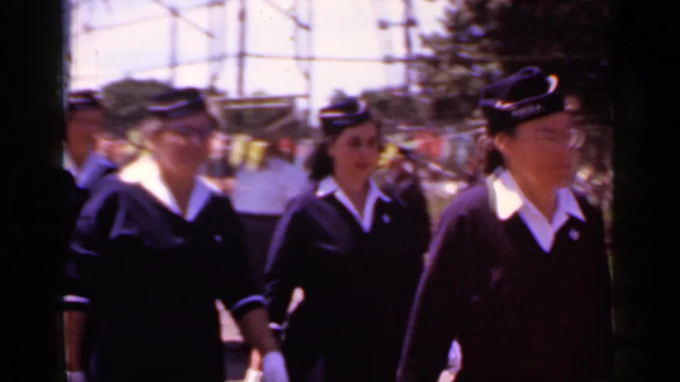 In 1966 Mineola, New York home-movie footage, women in dark uniforms with white collars and caps march along a residential street.