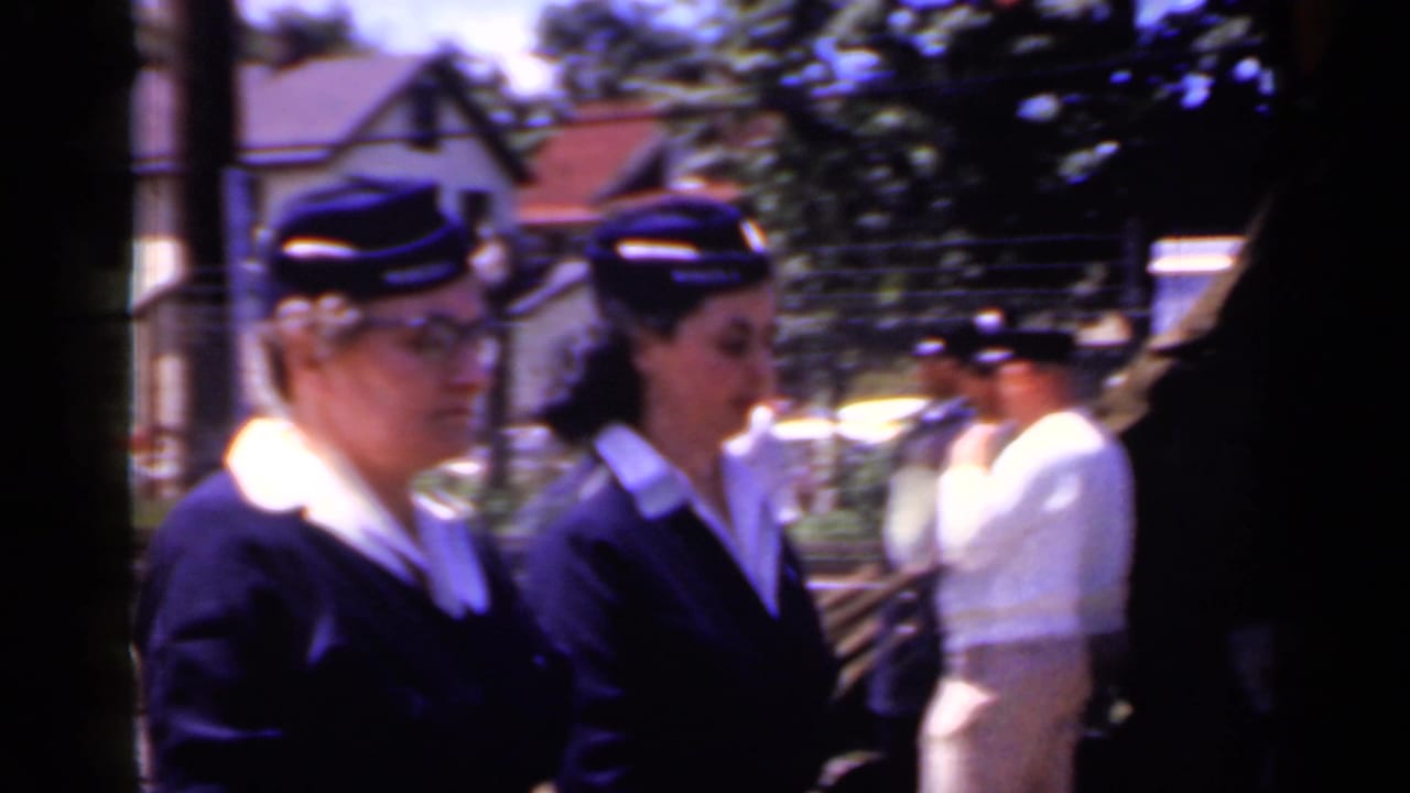 Close curbside view of women in matching dark parade uniforms and caps walking past in Mineola, New York, in 1966 footage.