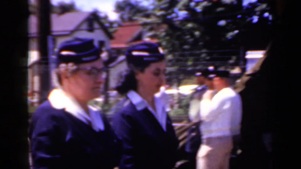 Close curbside view of women in matching dark parade uniforms and caps walking past in Mineola, New York, in 1966 footage.