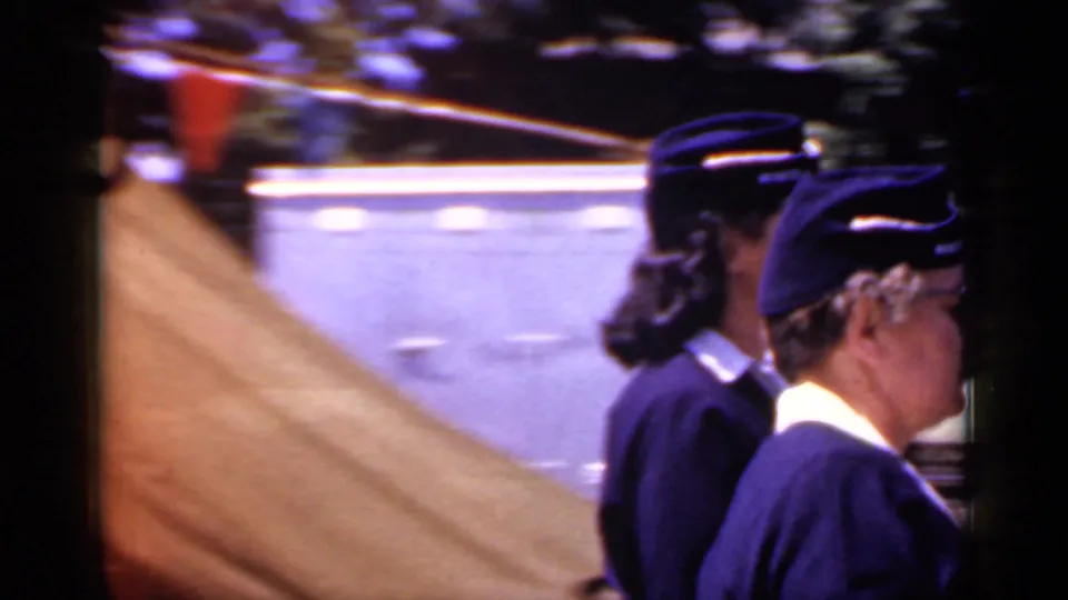 Side view of uniformed parade participants walking along a sunlit Mineola street, with the home-movie camera tracking from the curb.