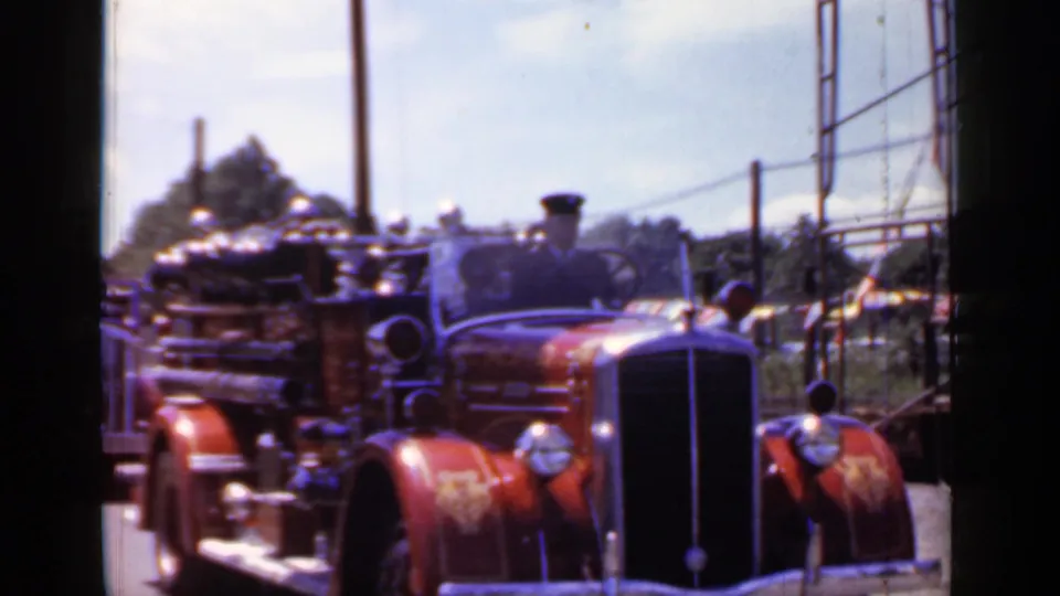 A red vintage firetruck appears in the 1966 Mineola parade footage, moving past fencing, poles, and spectators along the street.