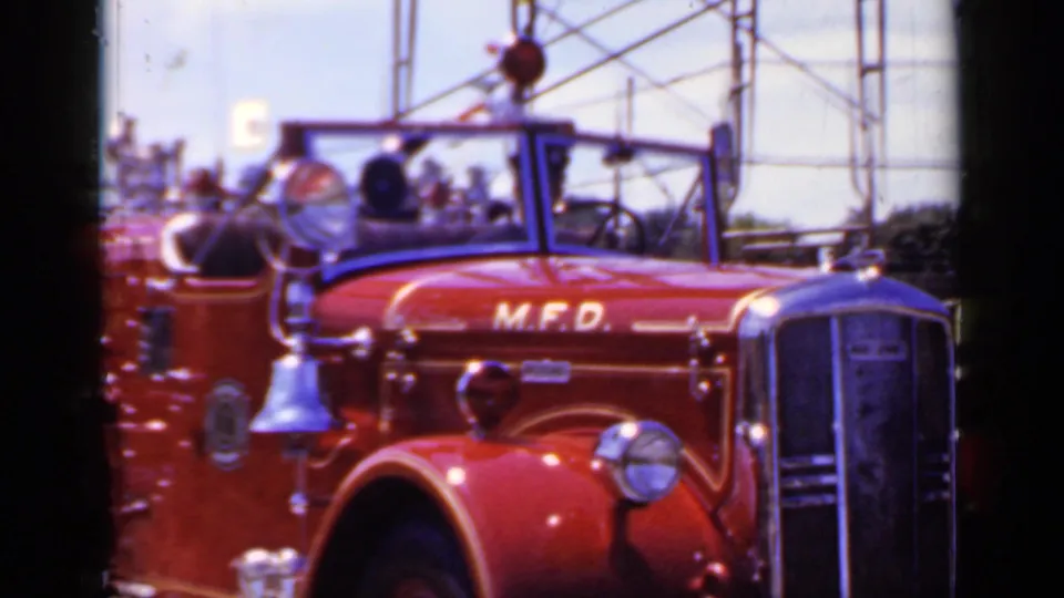 Close 1966 Mineola view of the red M.F.D. fire truck, emphasizing the hood lettering, side bell, chrome trim, and open cab.