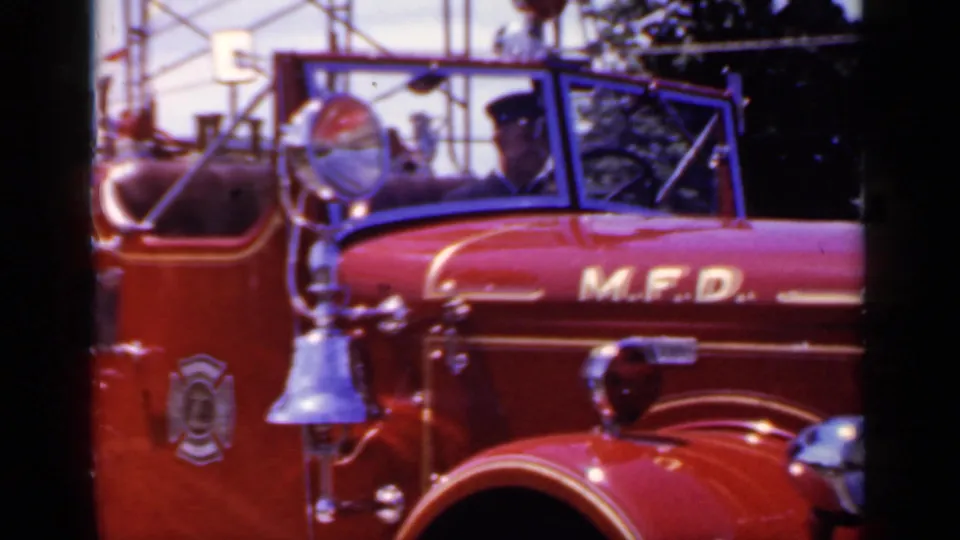 Red M.F.D. fire truck in Mineola with a uniformed driver visible through the open windshield and chrome bell mounted near the cab.