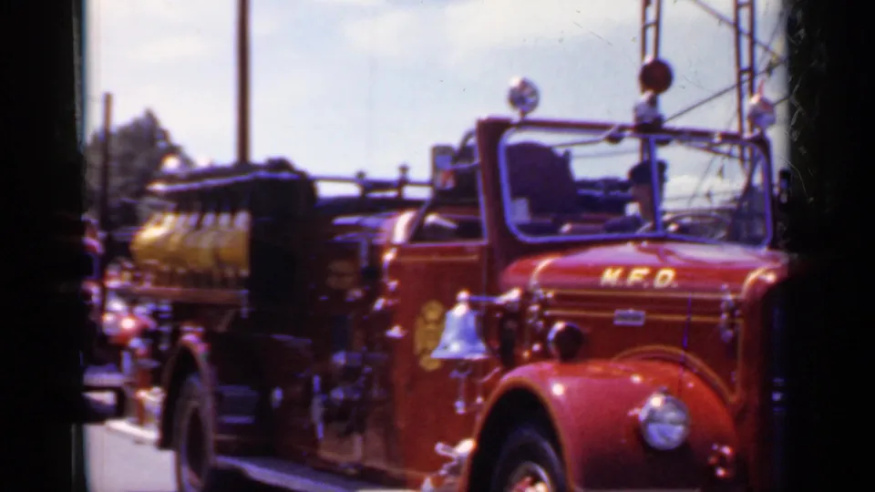 Front three-quarter view of the red M.F.D. fire truck during the 1966 Mineola parade, with bell, grille, and tank in view.