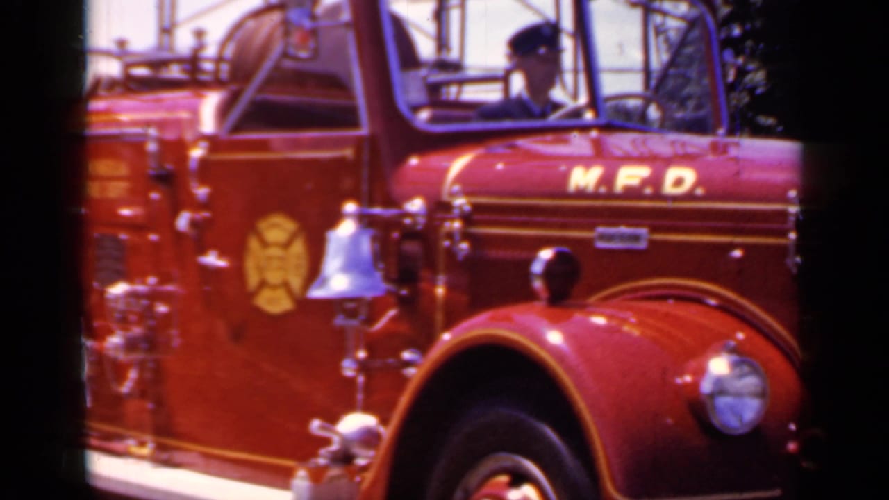 Red Mineola Fire Dept engine with M.F.D. lettering and a driver visible through the window.