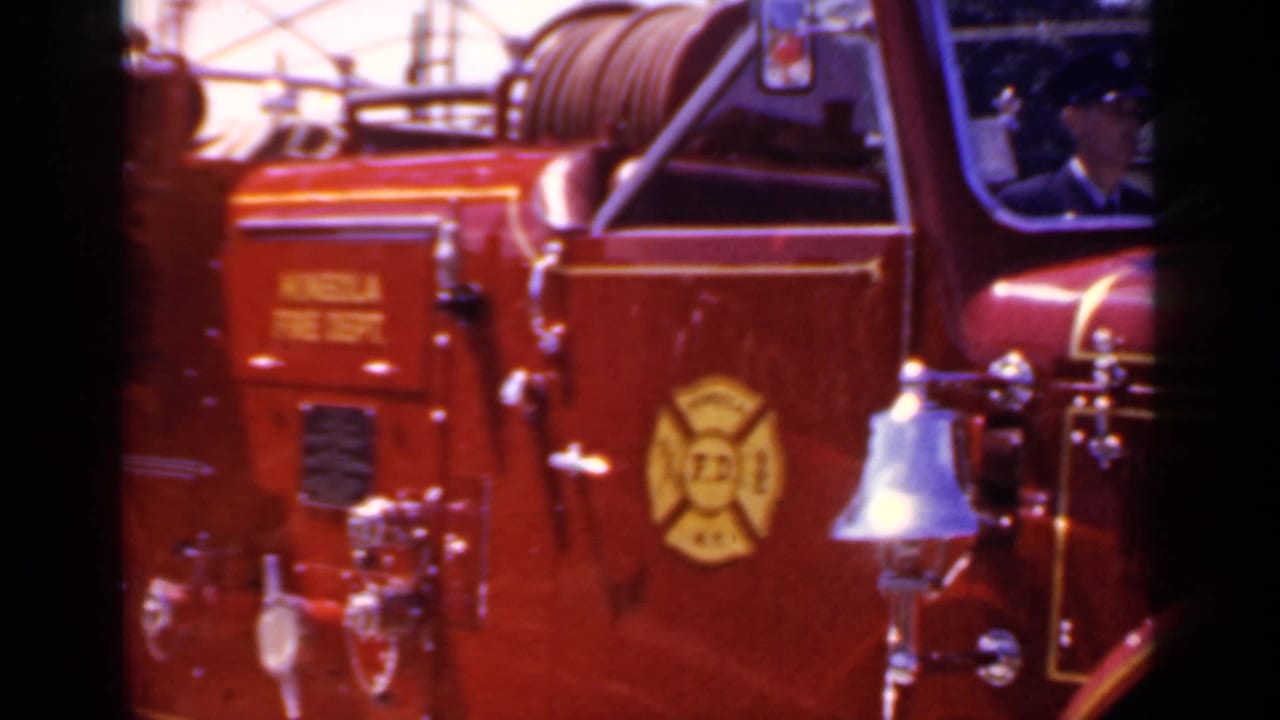 Side of a red Mineola Fire Dept engine with emblem, hose reel, and chrome bell.