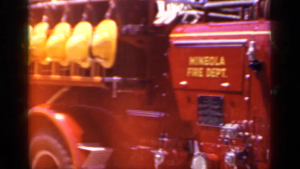 Home-movie frame of the Mineola Fire Dept engine side, with yellow helmets, red bodywork, department lettering, and chrome hardware visible.
