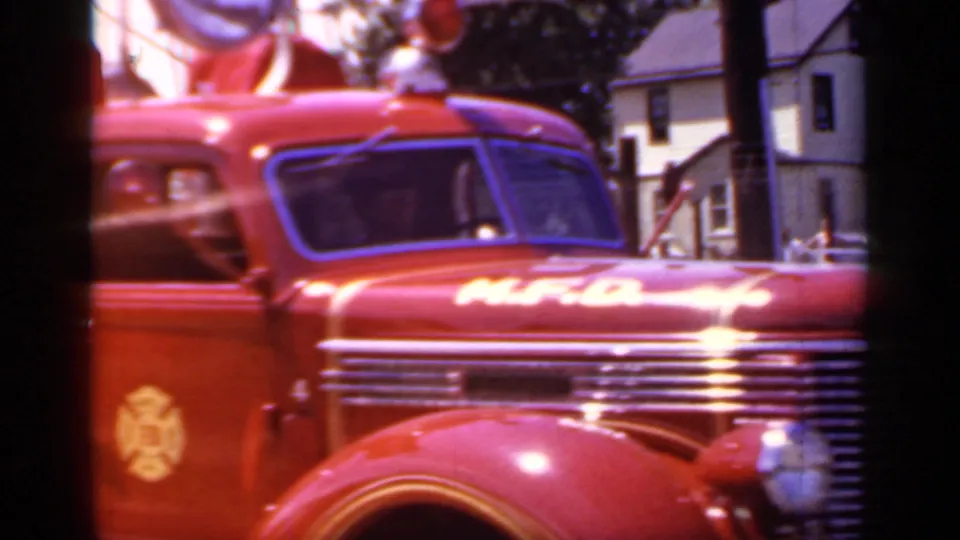 1966 Mineola, New York frame showing the red fire engine from the front side, with grille, cab window, emblem, and nearby houses visible.