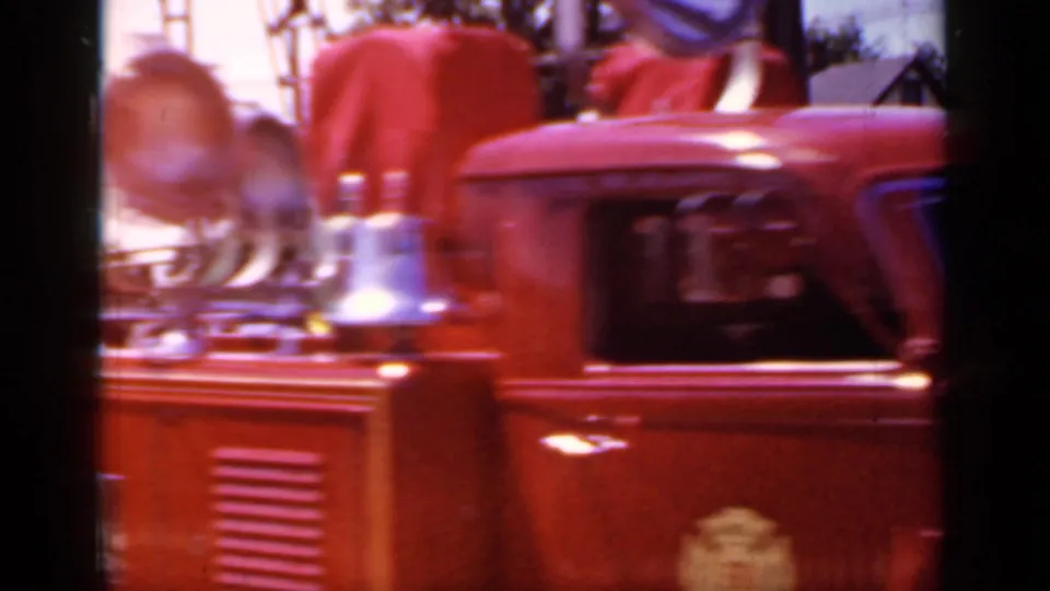 Close handheld 1966 Mineola parade view of a red fire truck body with chrome bell, hose fittings, and partial department insignia.