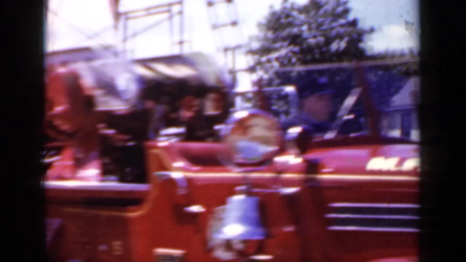 Close parade view of the red Mineola fire truck showing bell hardware, side lettering, chrome details, houses, trees, and overhead structure.