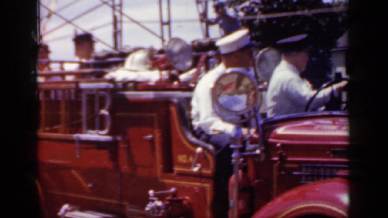 Uniformed firefighters seated on an open red fire truck during a parade.