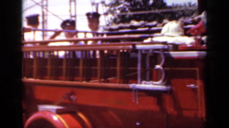 The side of the red parade fire truck fills the frame, with uniformed riders and equipment rails visible in the 1966 Mineola footage.