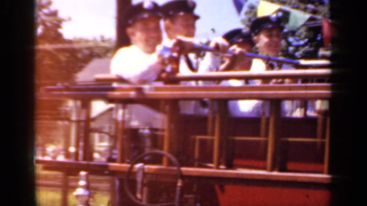 Firefighters in white shirts ride on an open red rescue vehicle during a parade.