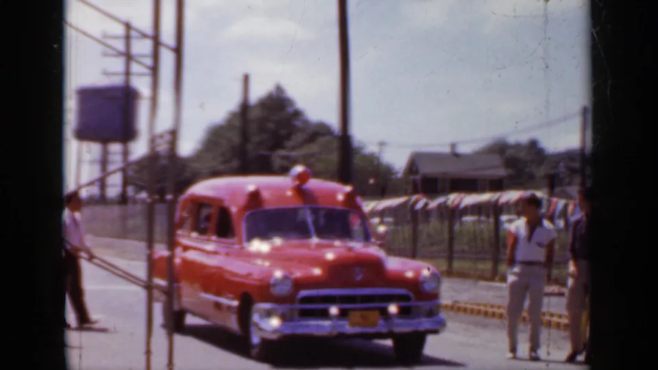 1966 Mineola street view with a red vintage rescue vehicle approaching past bunting and utility poles.