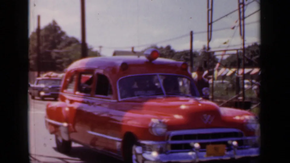 Red rescue vehicle in the 1966 Mineola parade passes roadside bunting, poles, trees, and parked cars.