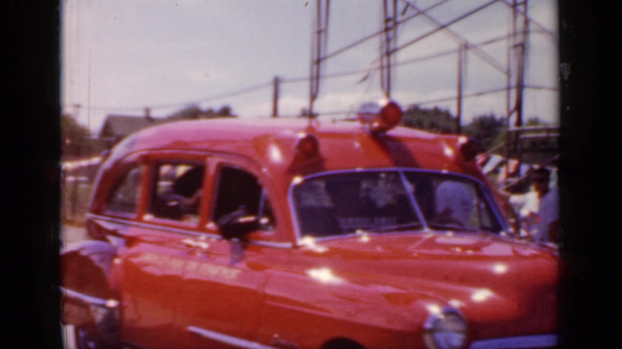 Red vintage ambulance driving past on a sunny street with people visible nearby.