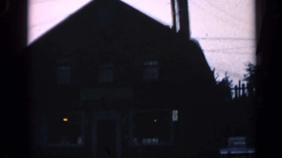 Shadowed 1968 Massachusetts 8mm view of the historic-looking building, showing lit windows, doorway, and a car beside the curb.