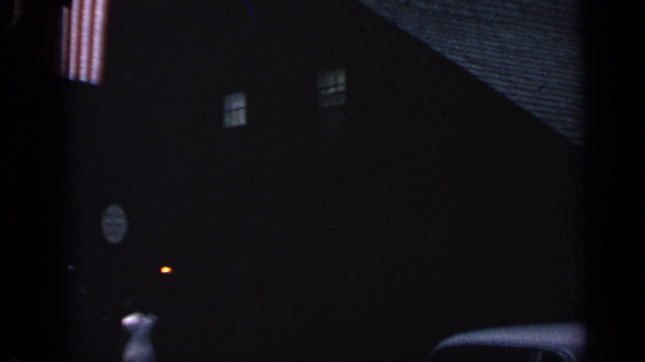 Woman in a light dress walking near a dark house, with windows and a small light visible.