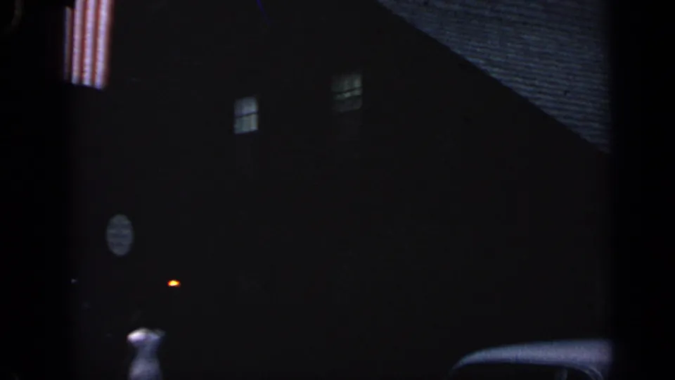 1968 Massachusetts home-movie frame with a woman in a white dress crossing in front of a dark two-story house at night.