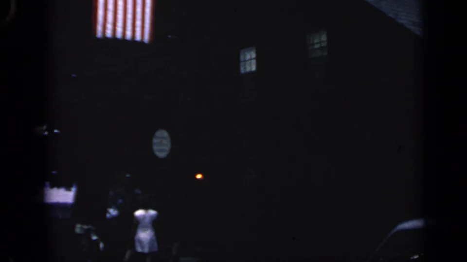 Nighttime 1968 Massachusetts 8mm frame with an American flag, dark house exterior, and woman in a white dress below.