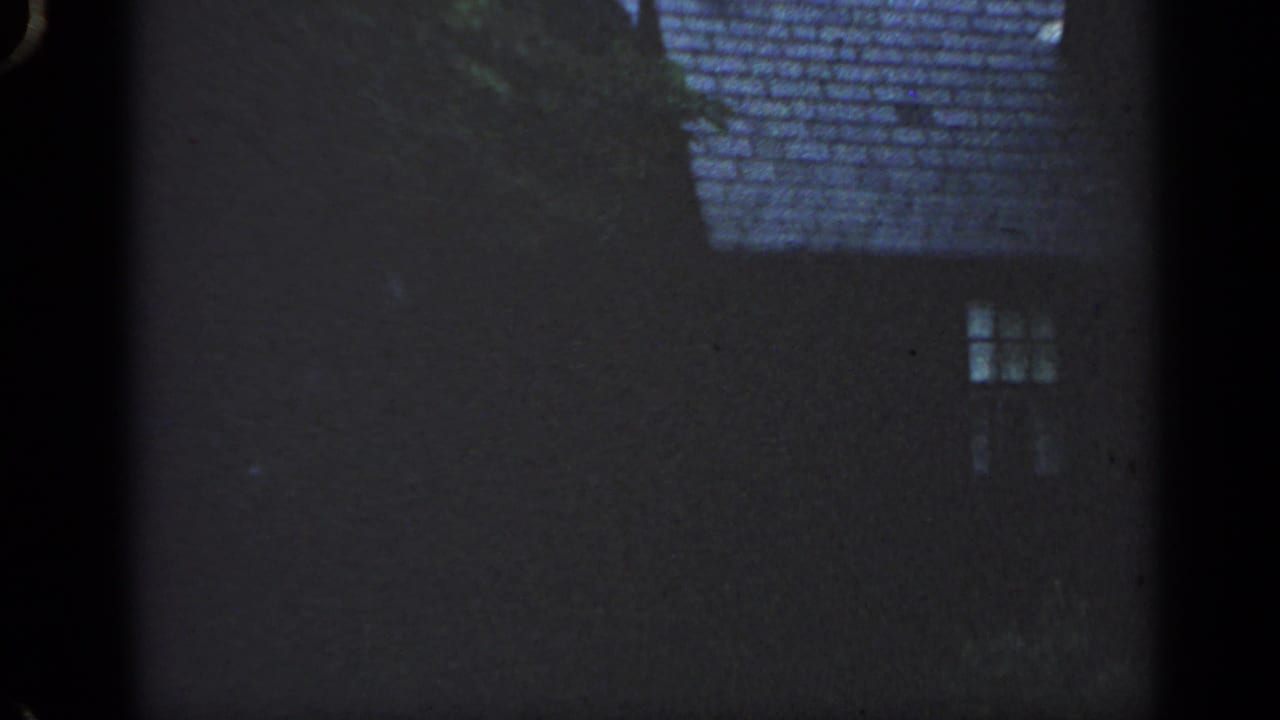1968 Massachusetts home-movie frame with a dark residential facade, shingled roof, small window, and tree cover against a pale sky.