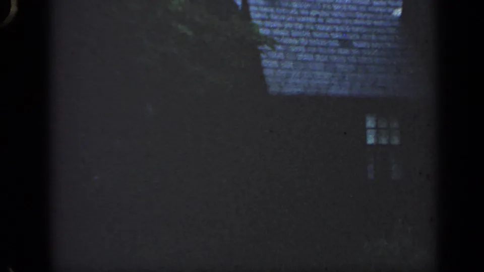 1968 Massachusetts home-movie frame with a dark residential facade, shingled roof, small window, and tree cover against a pale sky.