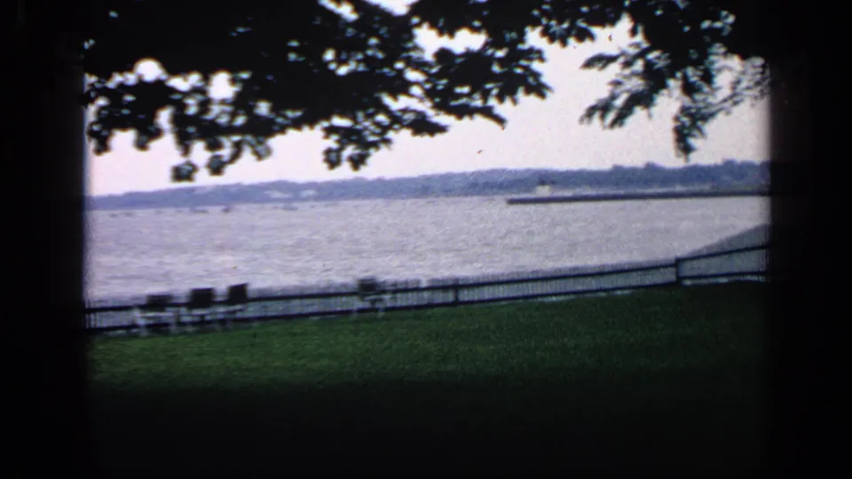 1968 Massachusetts home-movie frame showing a shaded park lawn, empty benches, low waterfront fence, lake, and distant shoreline.