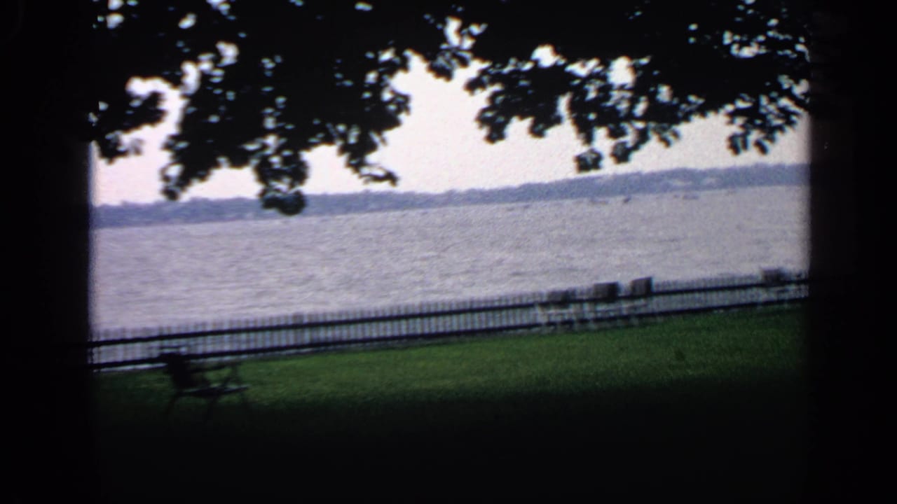 Waterfront park lawn seen through tree shade, with benches, fence, lake, and far shore.