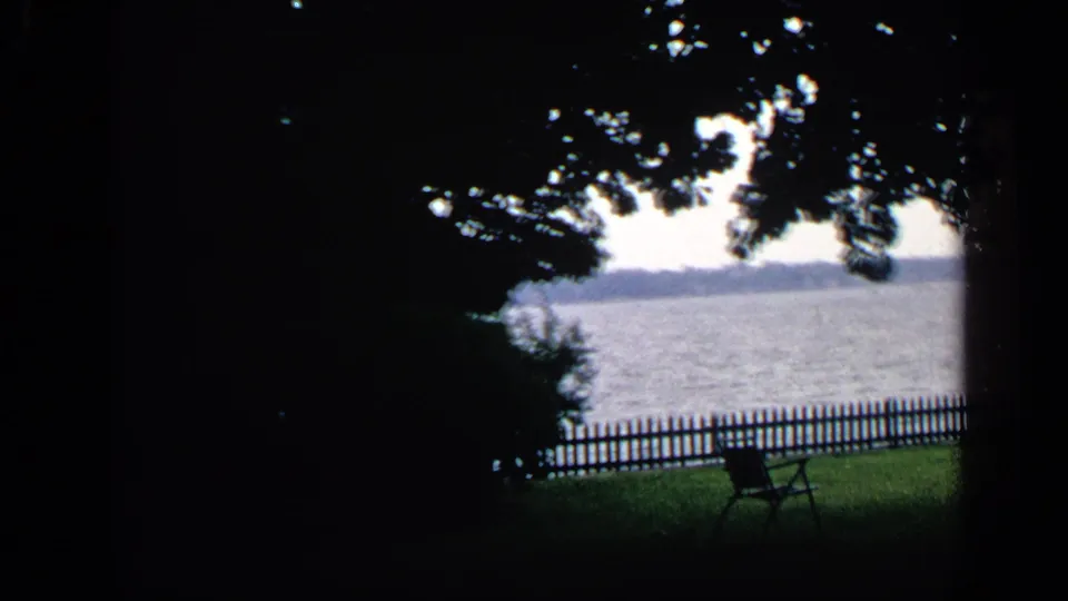 1968 Massachusetts home-movie view from shade toward a lakeside park bench, low fence, open water, and far shoreline.