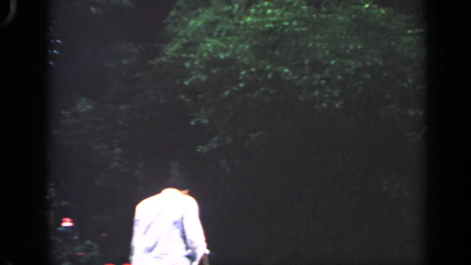 1968 Massachusetts 8mm frame showing a man in a light shirt walking through a dim yard near thick greenery.