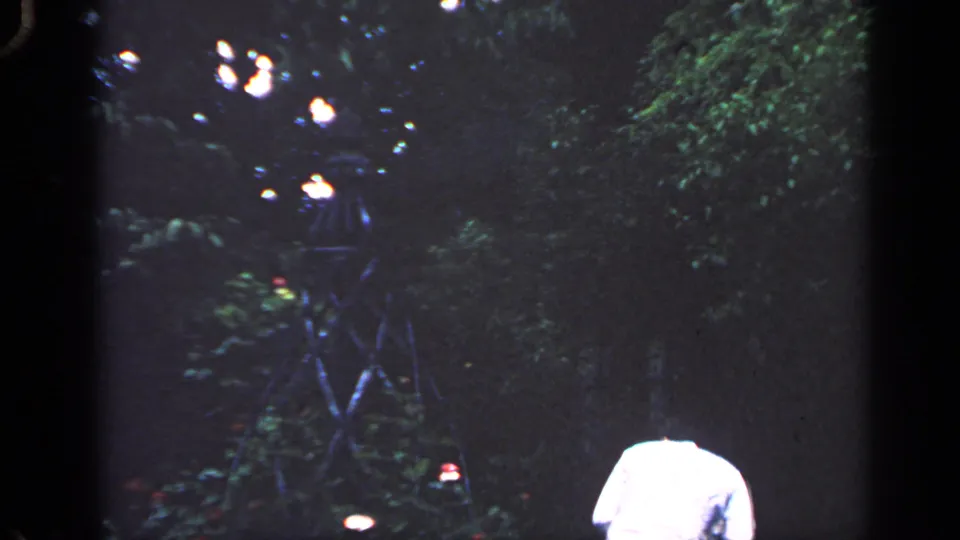1968 Massachusetts 8mm frame with a man near tree foliage and several small bright lights in a dark residential yard.