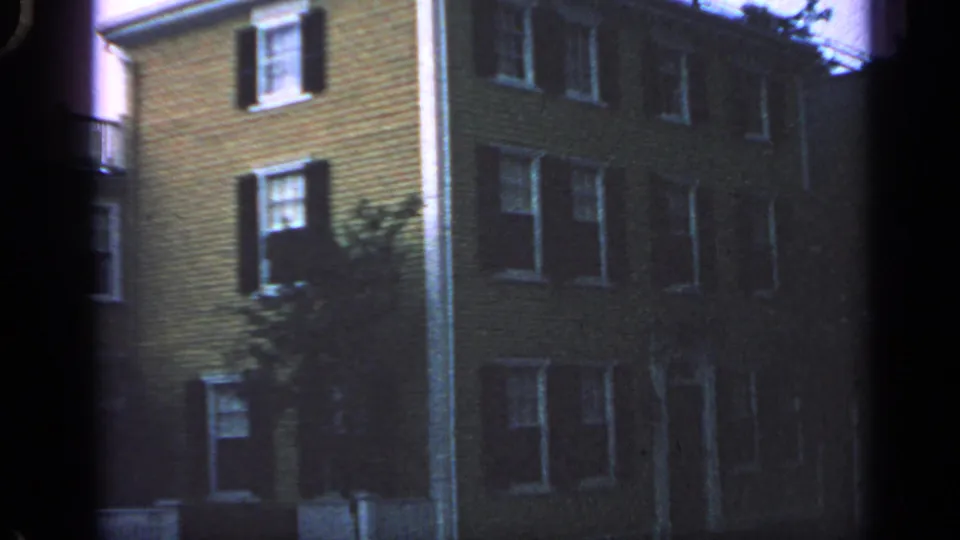 1968 Massachusetts 8mm frame showing a quiet residential block with shuttered windows, white trim, and a small tree beside the facade.