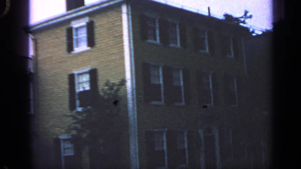 1968 Massachusetts home-movie still of a multi-story residential exterior with shuttered windows and a tree near the building side.