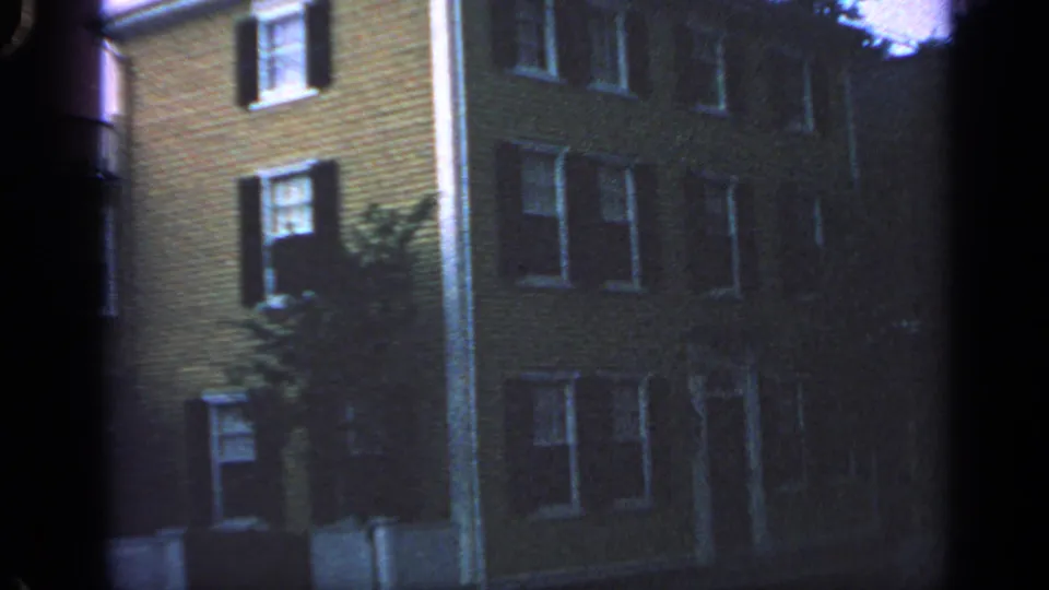 1968 Massachusetts home-movie frame of a quiet residential exterior with shuttered windows, a side tree, and visible film-edge vignetting.
