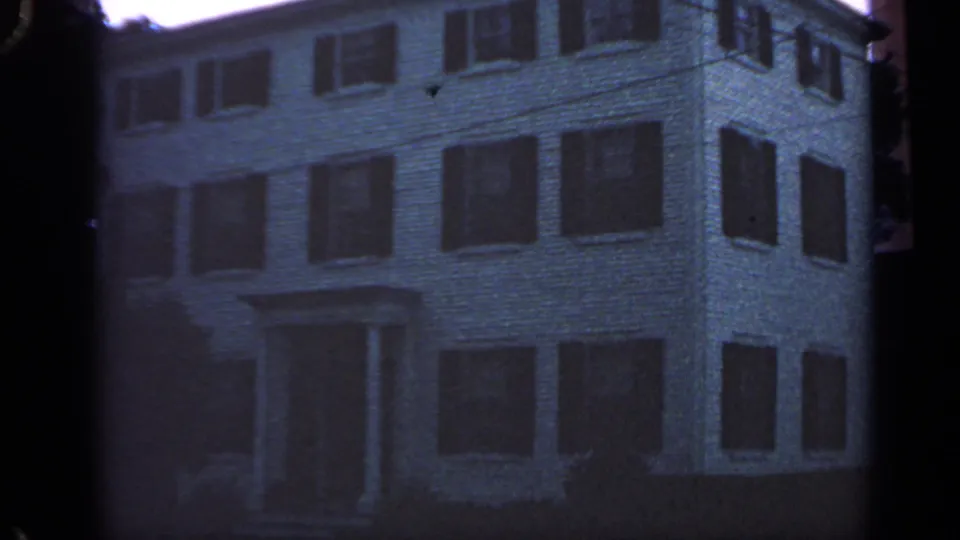 Handheld 1968 Massachusetts frame showing the building corner, rows of shutters, and entrance area in dim exposure.